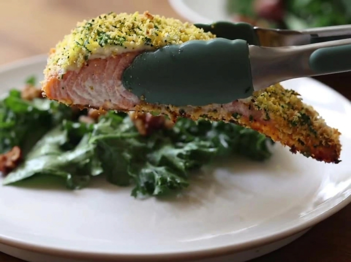 Pinzas de cocina de silicona verde sosteniendo un trozo de salmón horneado con una costra dorada y crujiente sobre un plato blanco que contiene una ensalada de col rizada verde.