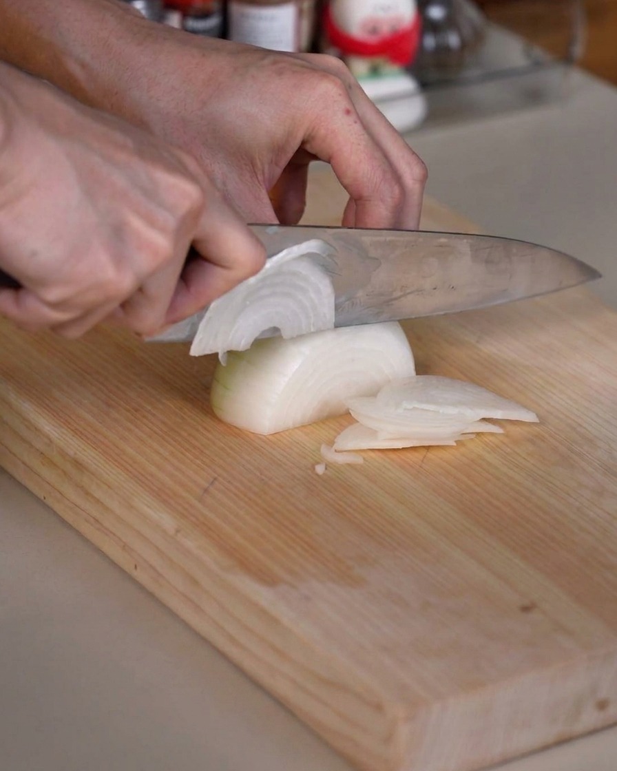 Una cebolla cortándose cuidadosamente en tiras finas sobre una tabla de picar de madera con un cuchillo de cocina afilado.