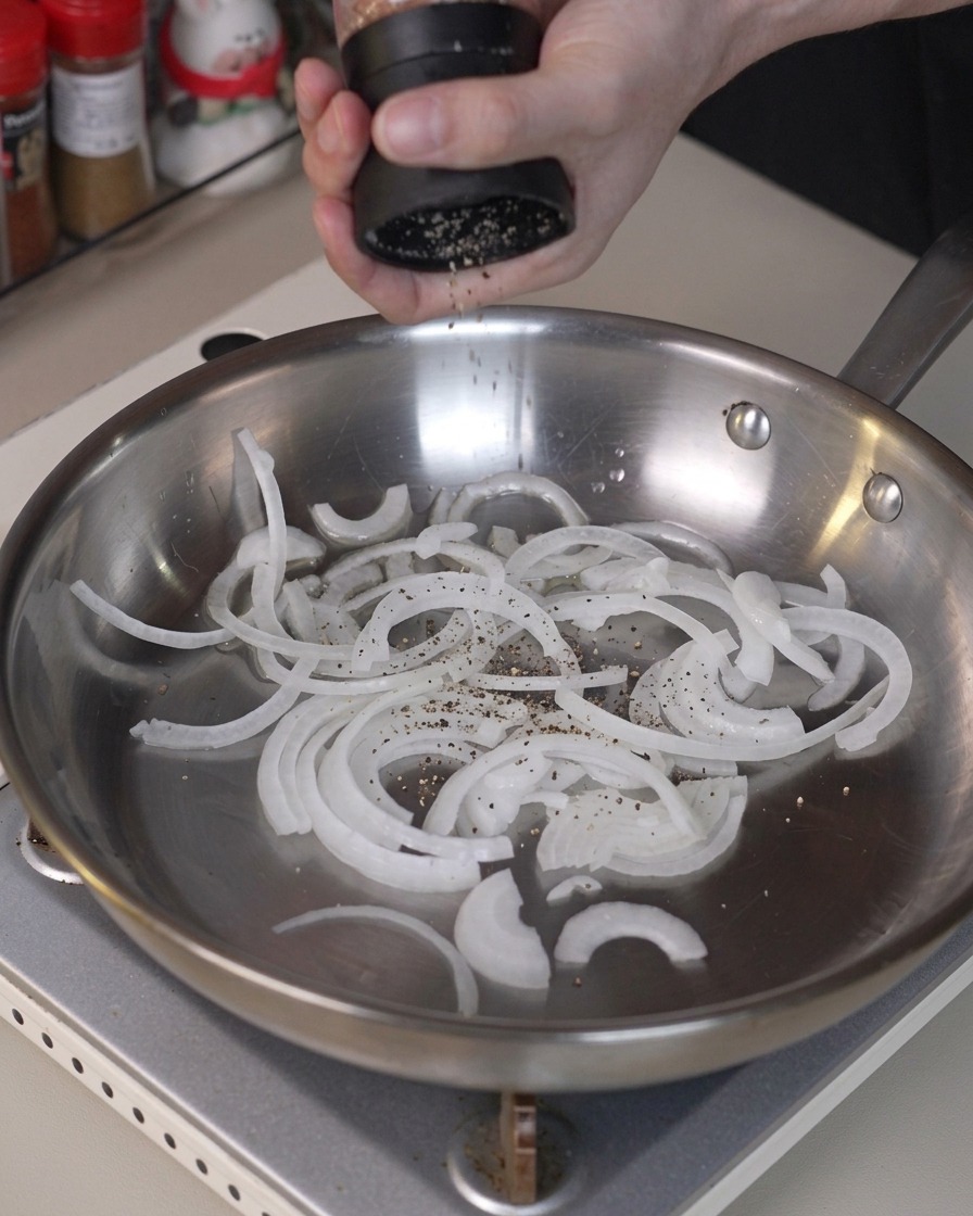 Cebollas en rodajas sazonadas con pimienta y salteadas en una sartén de acero inoxidable.