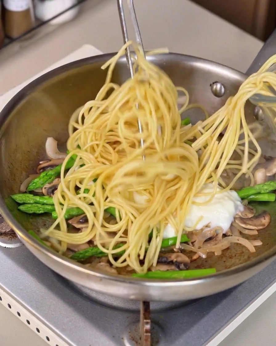 Pasta espagueti larga levantada con pinzas y mezclada en la sartén con salsa blanca y verduras.