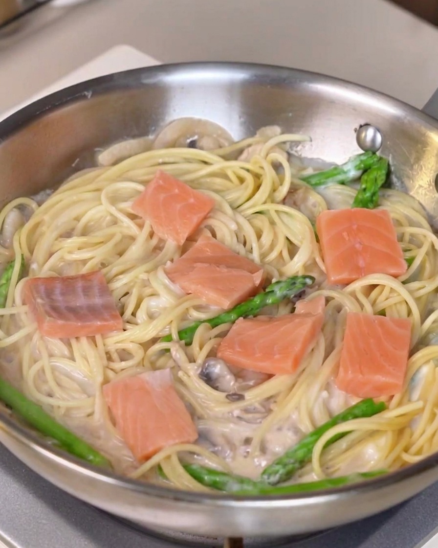 Trozos frescos de salmón distribuidos sobre una sartén de pasta cremosa y espárragos.