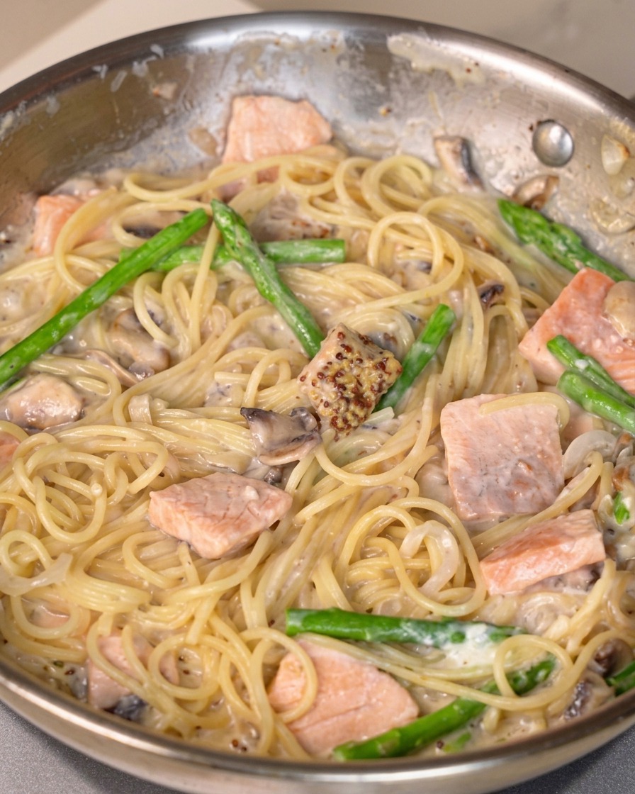 Salsa de mostaza, sal y pimienta mezclándose en una sartén de pasta cremosa con salmón.