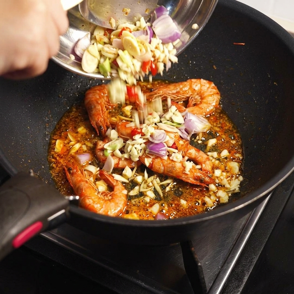 Aromáticos picados y pasta Tom Yum agregados desde un bol a una sartén con cáscaras de langostino fritas.