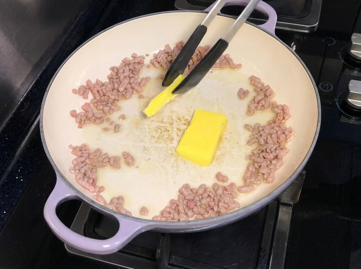 Mantequilla siendo agregada y derretida en una sartén con cerdo picado dorado.
