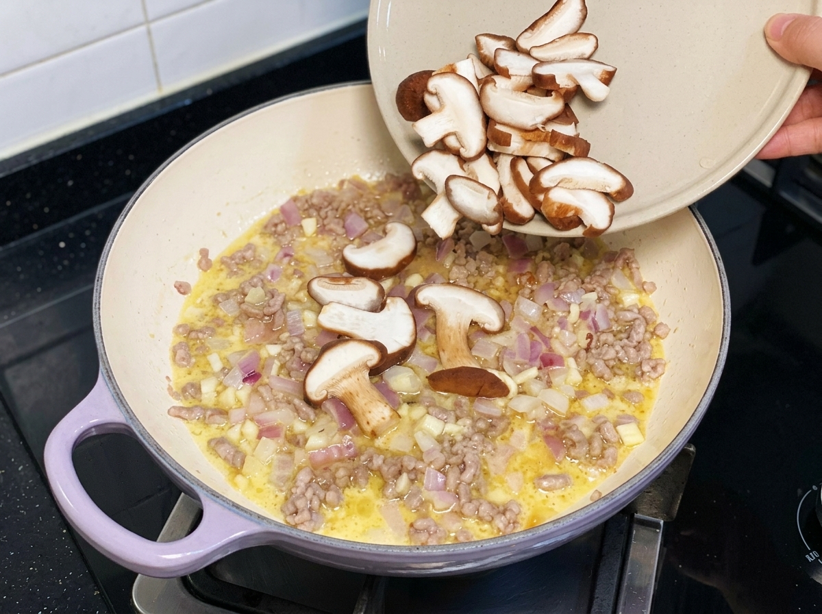Champiñones shiitake frescos y rebanados siendo vertidos desde un plato hacia una sartén con cerdo picado y cebollas.