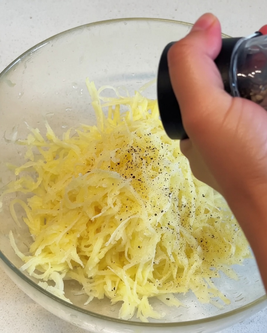 Patatas amarillas ralladas dentro de un bol de cristal siendo sazonadas con pimienta negra de un molinillo manual.