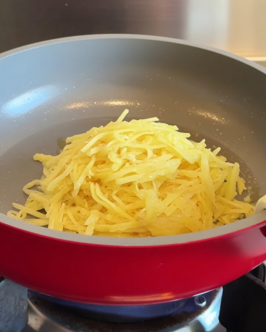 Una pila de patatas ralladas colocada en el centro de una sartén antiadherente roja caliente.