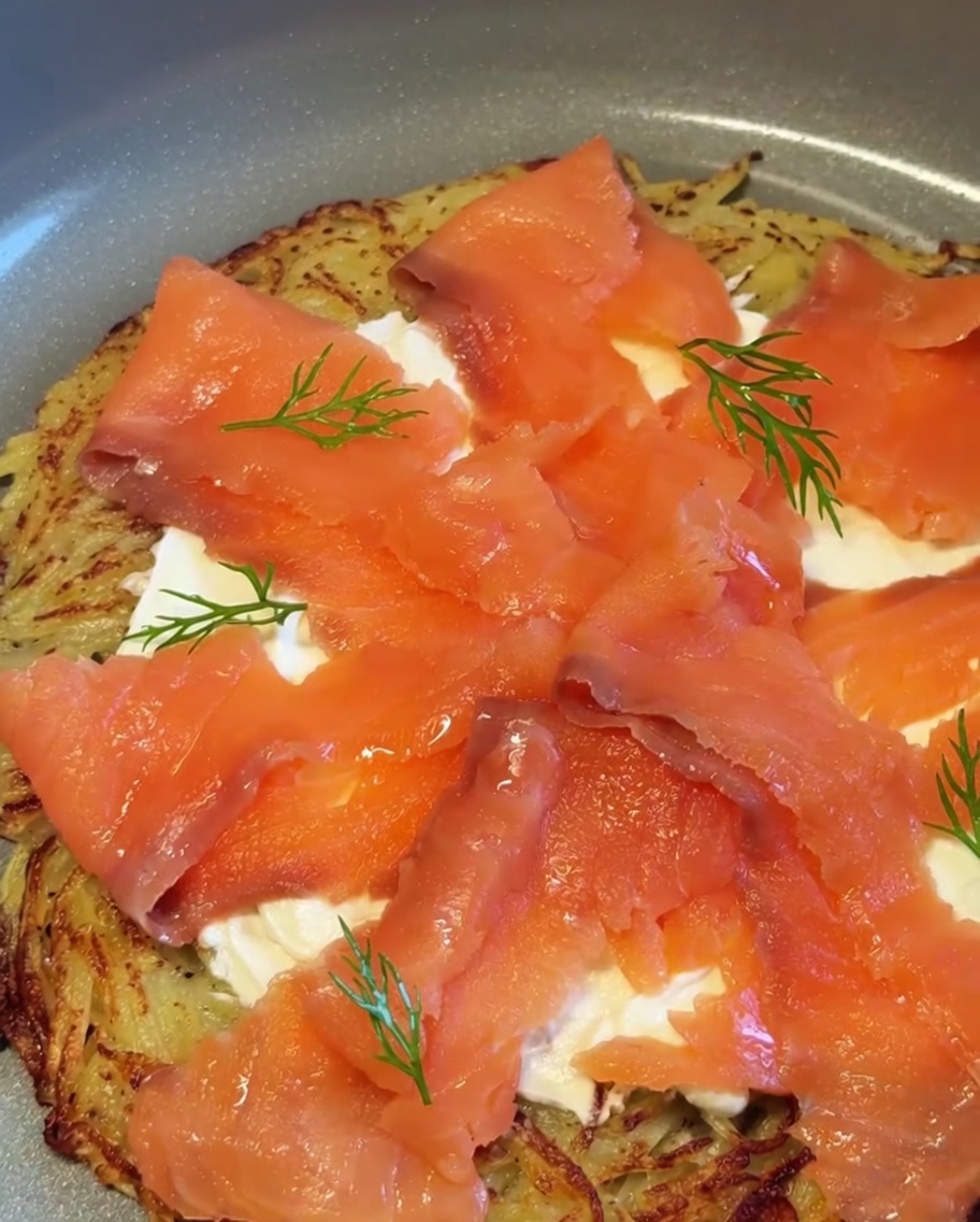 Una pizza terminada de patata y salmón ahumado adornada con ramitas de eneldo fresco y trozos de cebolla morada.