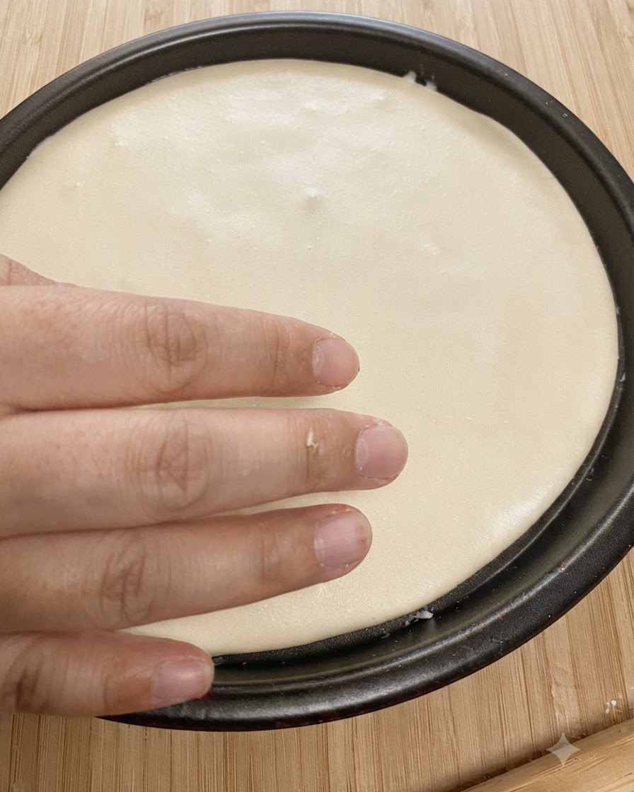 Masa redonda aplanada colocada dentro de un molde negro para hornear con una mano presionando los bordes.