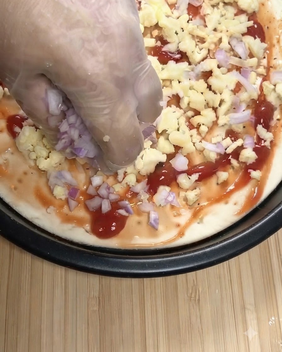 Una mano con un guante transparente esparciendo cebollas moradas picadas y mozzarella rallada sobre una base de pizza con salsa de tomate.