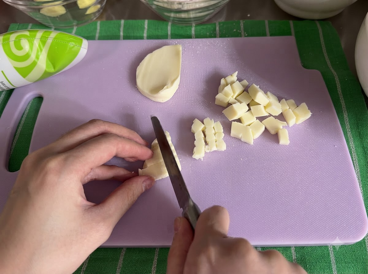 Un bloque de queso mozzarella siendo cortado en cubos pequeños con un cuchillo sobre una tabla de cortar.
