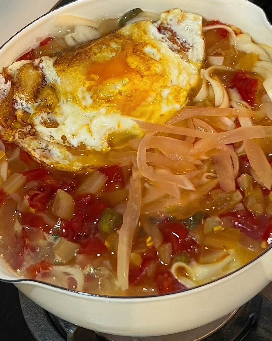 Cilantro fresco picado siendo esparcido sobre una olla burbujeante de fideos, repollo encurtido y un huevo frito.