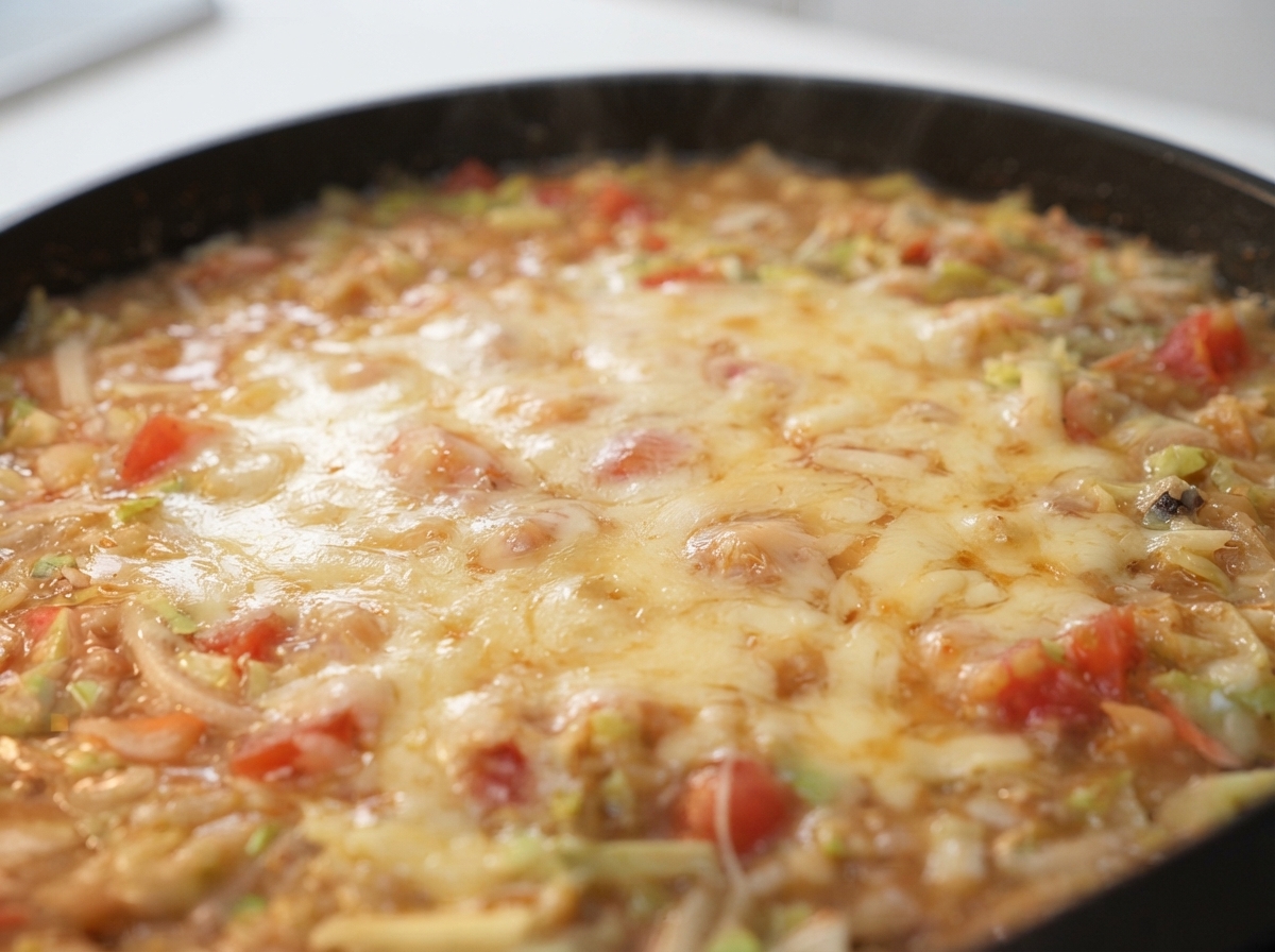 Queso rallado derritiéndose sobre una mezcla burbujeante de masa y verduras en una sartén caliente.