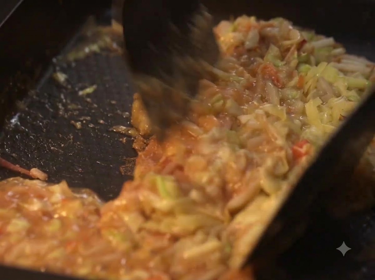 Espátulas mezclando verduras cocinadas y masa líquida en una placa caliente.
