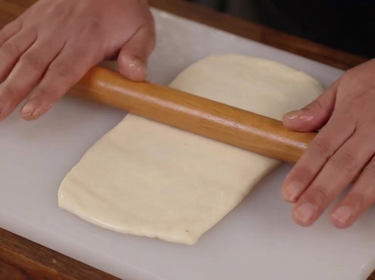 Manos usando un rodillo de madera para aplanar una pieza de masa lisa en forma de rectángulo sobre una tabla de cortar blanca.