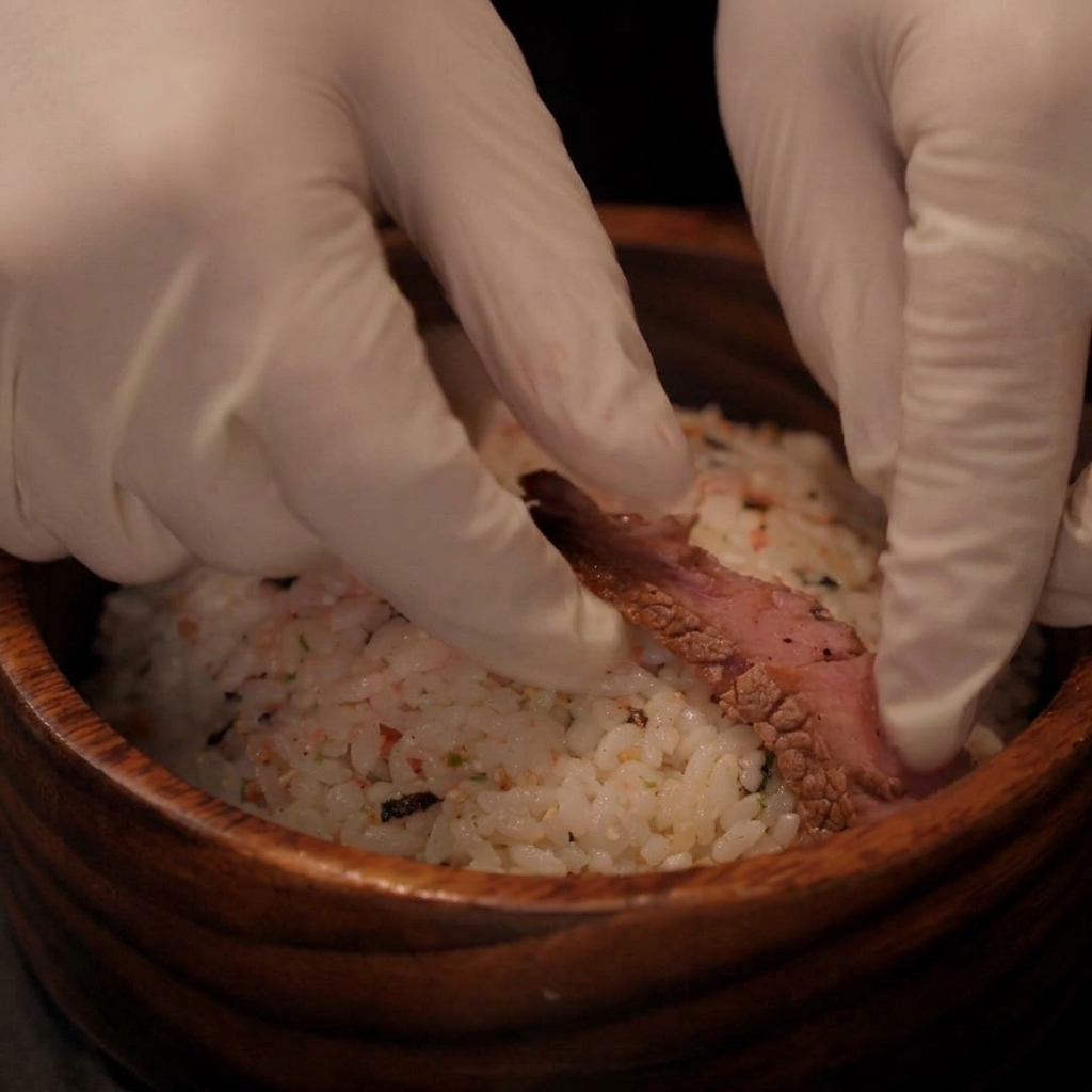 Manos con guantes colocando rebanadas de carne cocida sobre un tazón de arroz sazonado.