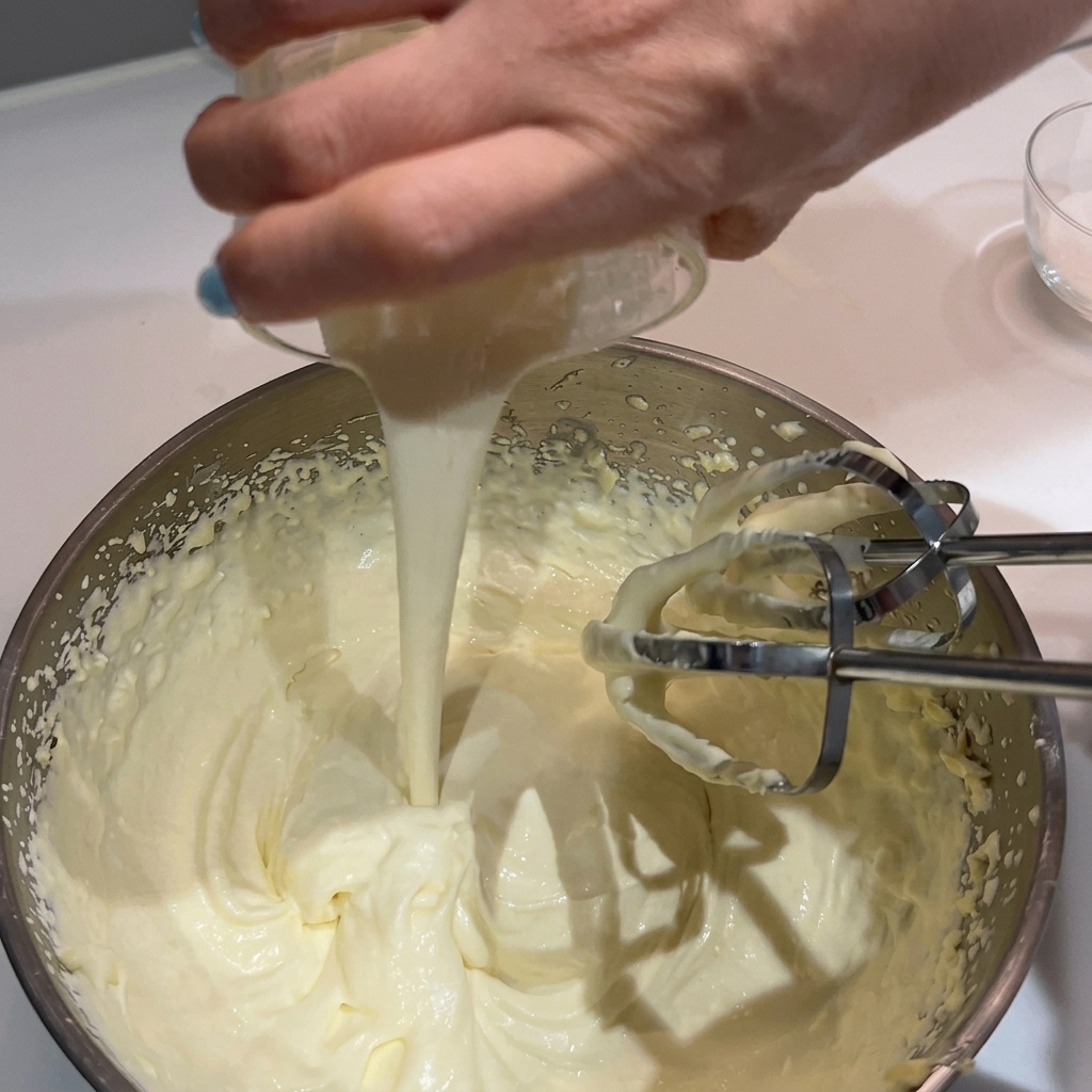 Una mano vertiendo más nata en un bol de nata montada y mantequilla con varillas eléctricas.