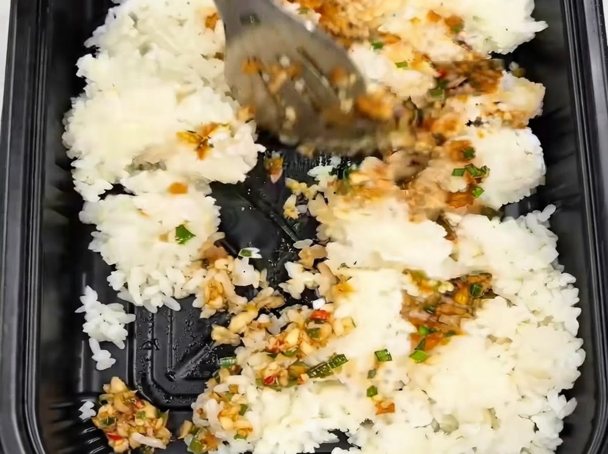 Arroz siendo revuelto y mezclado con pinzas dentro de un recipiente negro para llevar junto con la salsa de ajo.