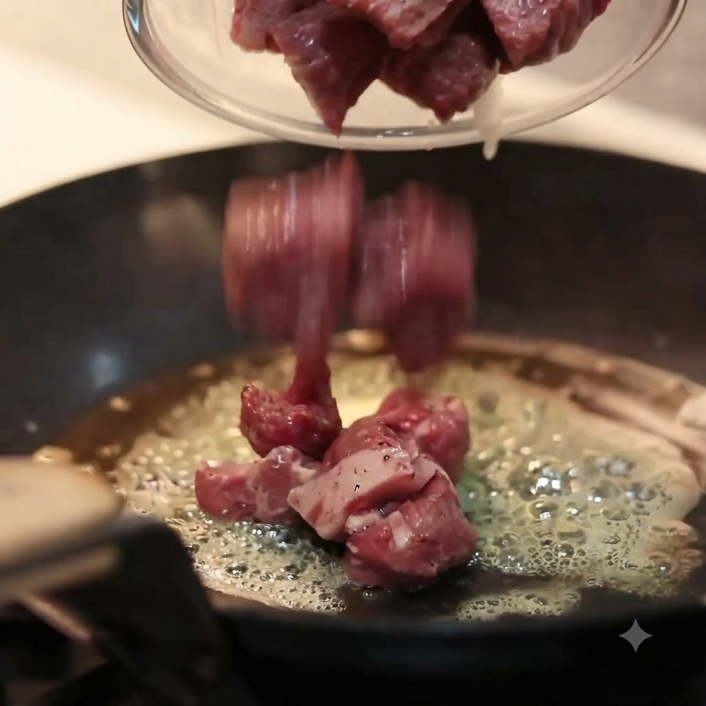 Trozos de carne cruda sazonada vertiéndose desde un bol de cristal a una sartén caliente de fondo plano con mantequilla derretida burbujeante.