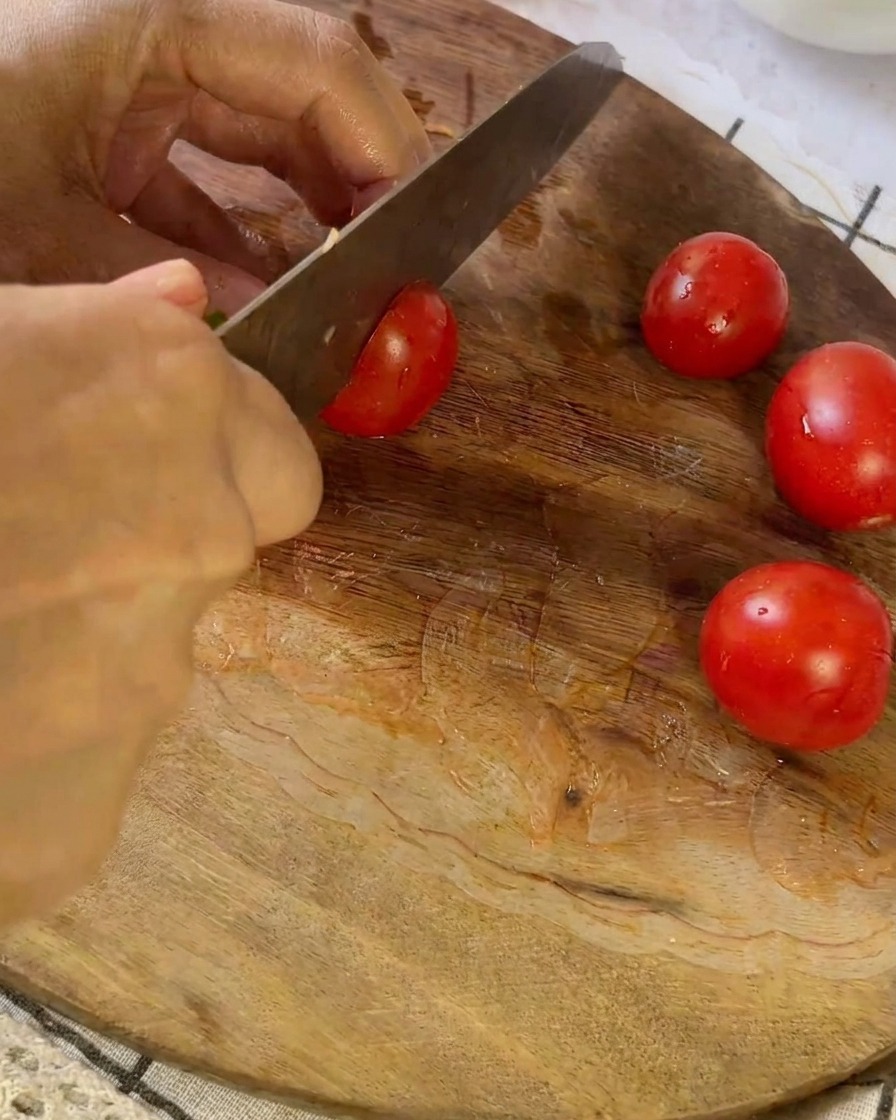 Un cuchillo cortando un pequeño tomate rojo en una tabla de cortar de madera junto a otros tomates cortados.