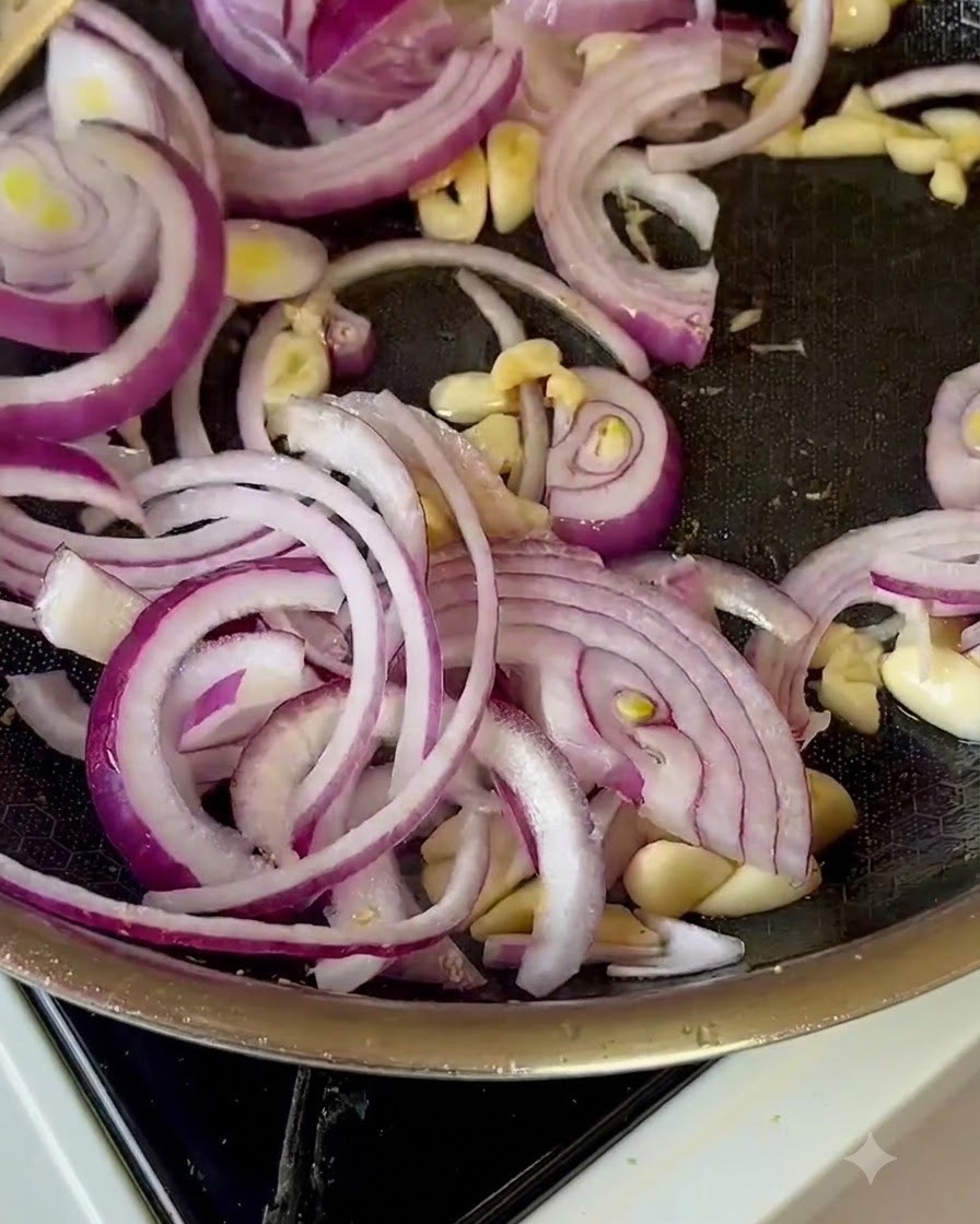 Cebollas moradas en rodajas, ajo y trozos de manzana siendo revueltos en una sartén.