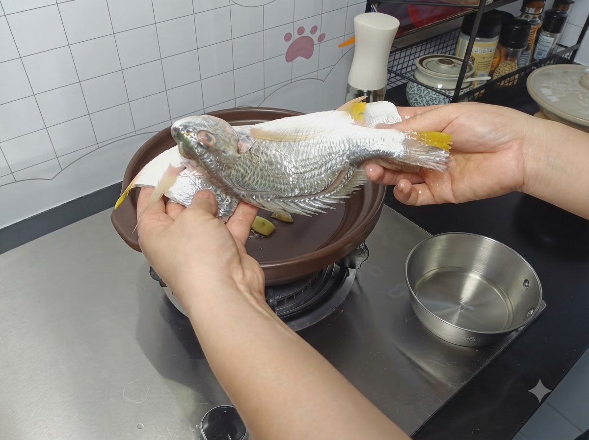 Un pescado entero sazonado siendo colocado en una cazuela de barro sobre rodajas de jengibre.