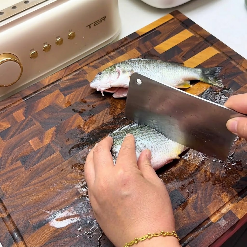 Una mano utilizando un cuchillo grande para hacer cortes diagonales en el costado de un pescado fresco sobre una tabla de cortar de madera.
