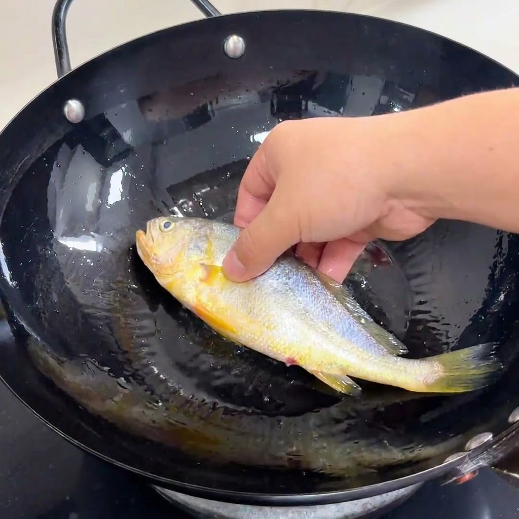 Una mano colocando suavemente un pescado entero sazonado en un wok negro lleno de aceite de cocina caliente.