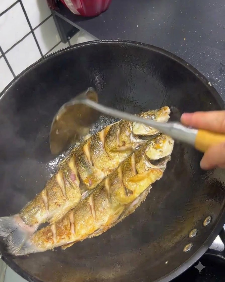 Dos pescados de cola amarilla friéndose en un wok con la piel dorada siendo manipulados con una espátula.