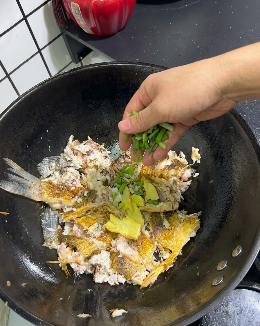 Jengibre fresco en rodajas y cebollas verdes picadas siendo añadidas a mano sobre el pescado triturado en un wok.