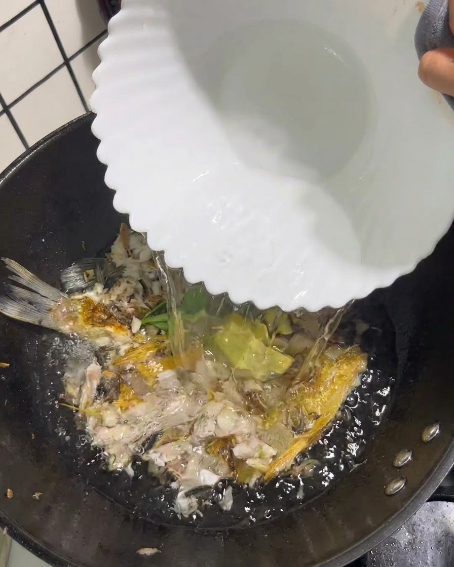 Agua hirviendo y clara siendo vertida desde un tazón blanco en un wok que contiene pescado triturado y aromáticos.