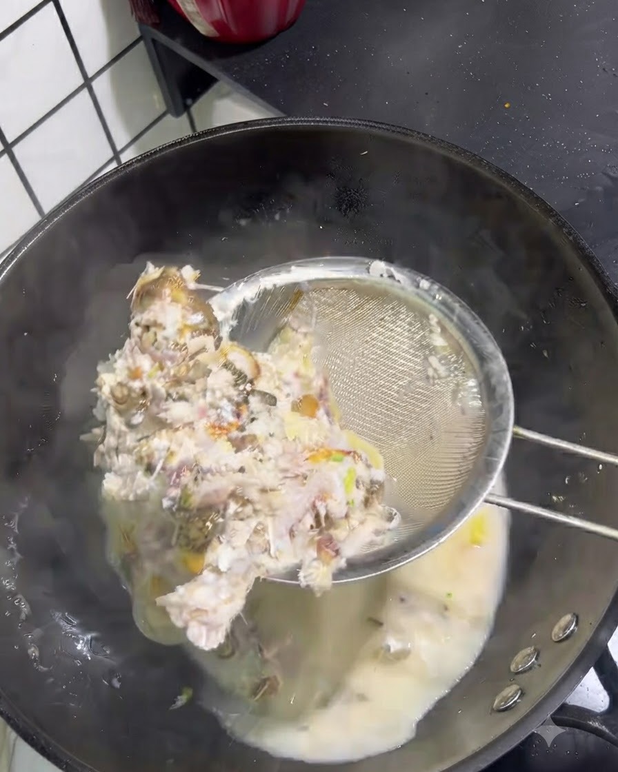 Espinas y carne de pescado sólido siendo retiradas de un caldo lechoso con un colador de malla metálica.