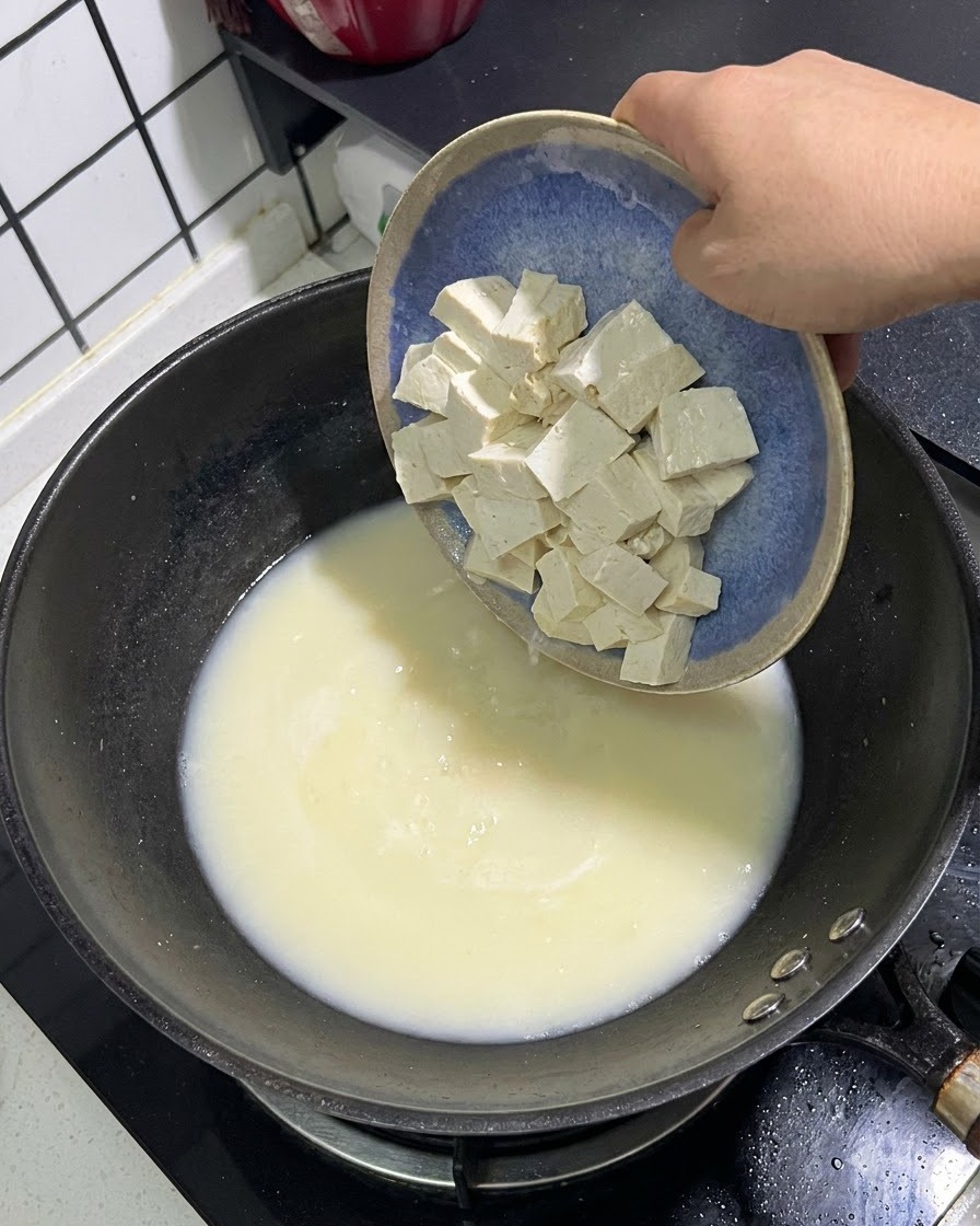 Cubos de tofu blanco deslizándose desde un plato azul hacia una olla de caldo de pescado blanco cremoso.
