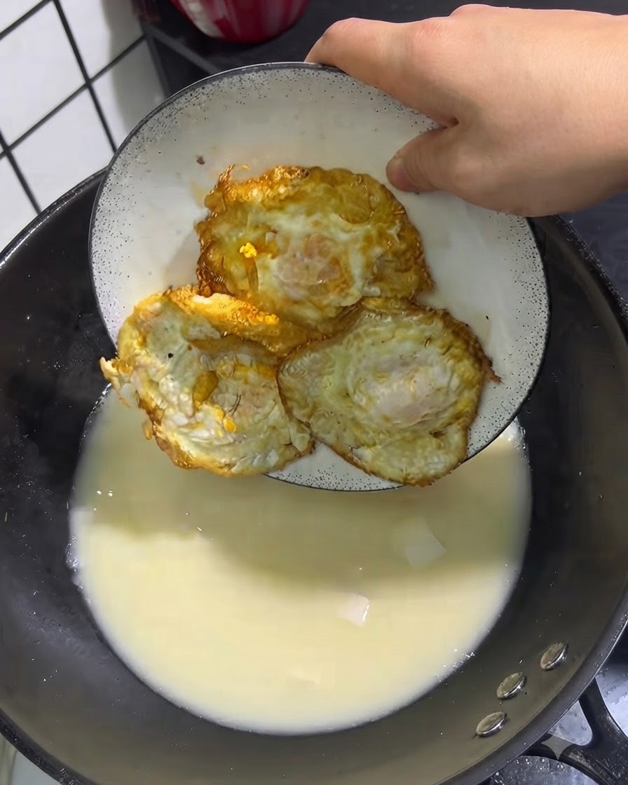 Tres huevos fritos dorados siendo colocados desde un plato blanco en una olla de sopa a fuego lento.