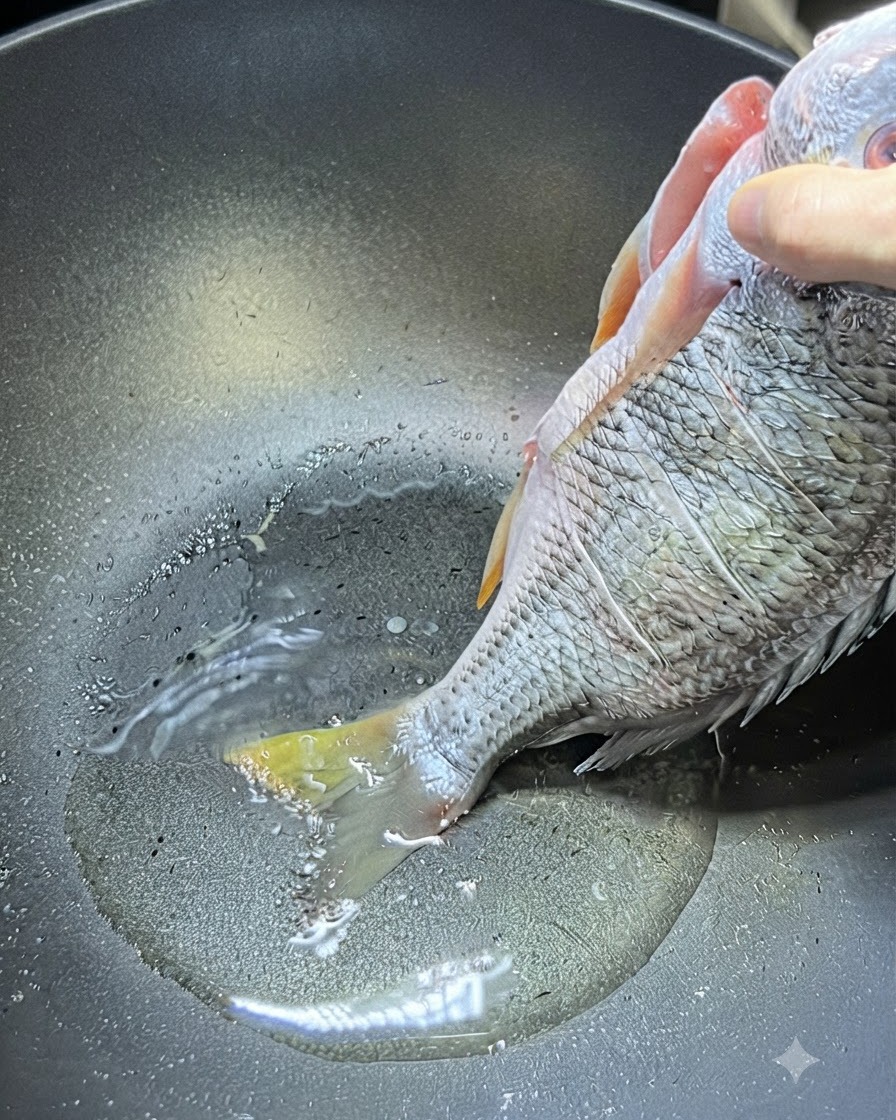 Una mano sosteniendo un pargo amarillo crudo entero por la parte superior del cuerpo mientras la cola toca el aceite caliente en un wok.