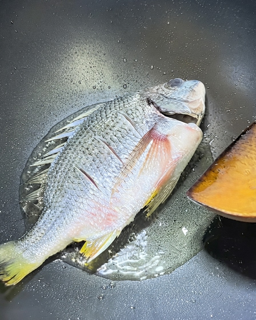 Un pargo amarillo entero friéndose en un wok con una espátula de madera cerca lista para voltearlo.