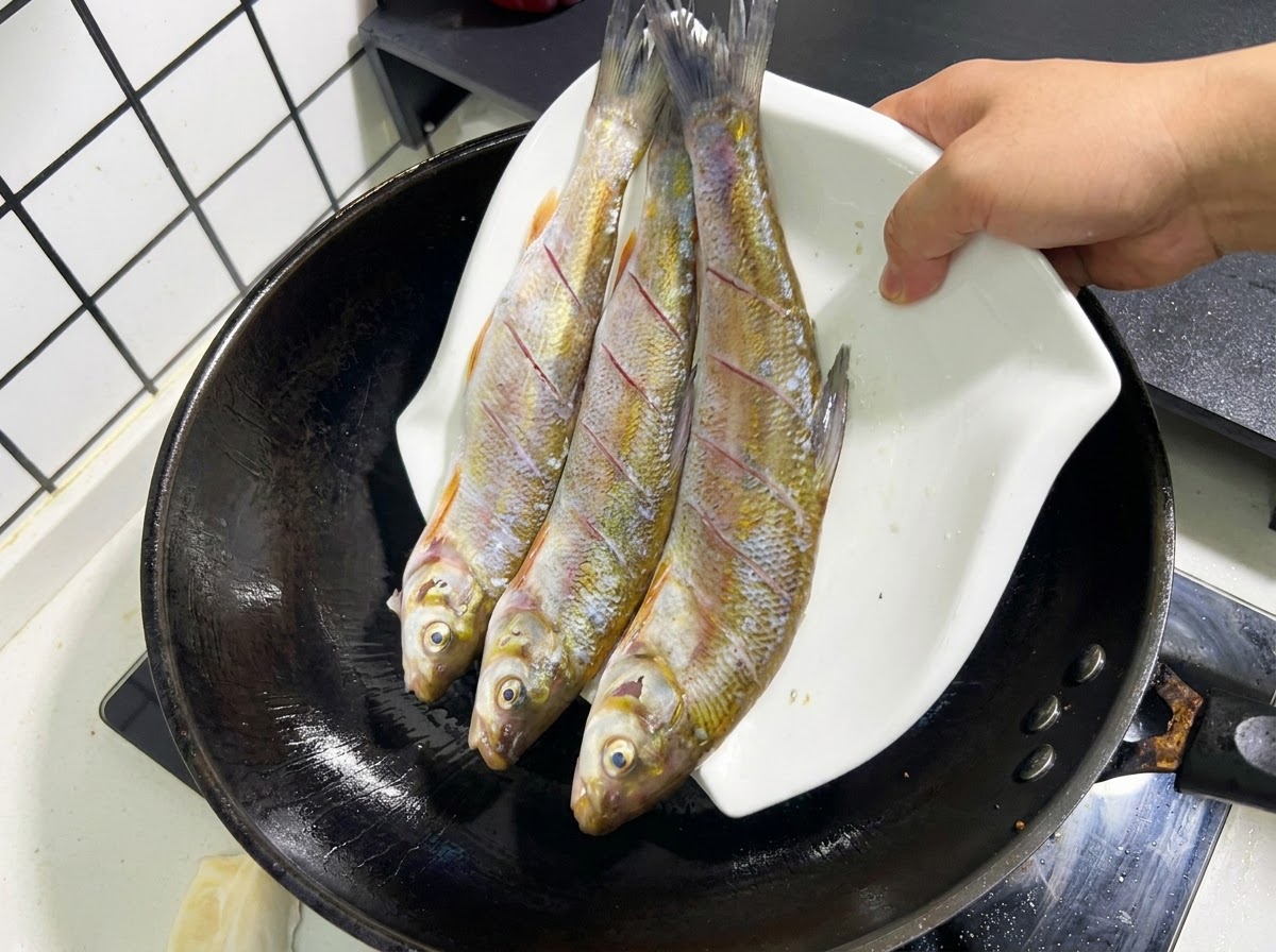 Tres pescados enteros crudos con cortes siendo deslizados desde un plato blanco hacia un wok negro con aceite caliente.