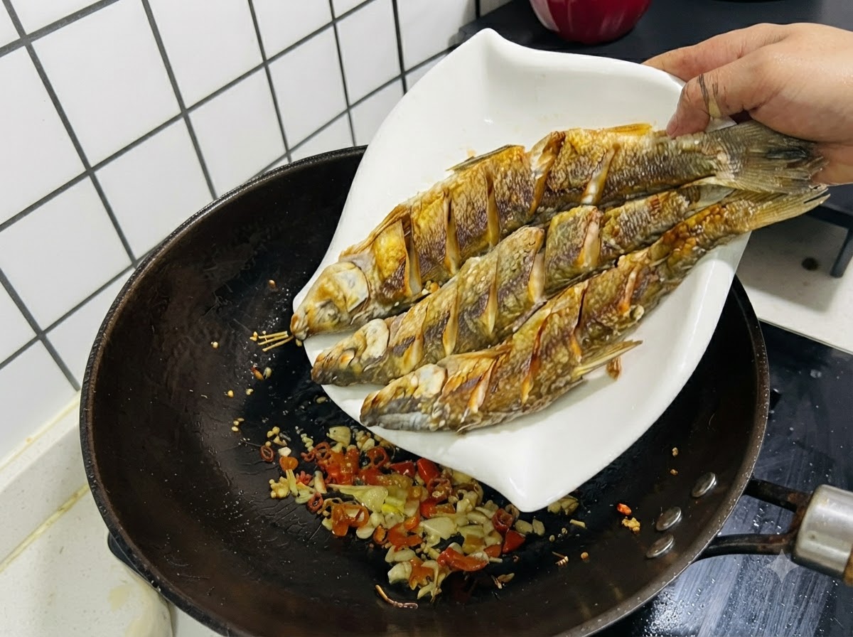 Tres pescados fritos siendo deslizados desde un plato blanco de regreso a un wok que contiene ajo, jengibre y chiles salteados.