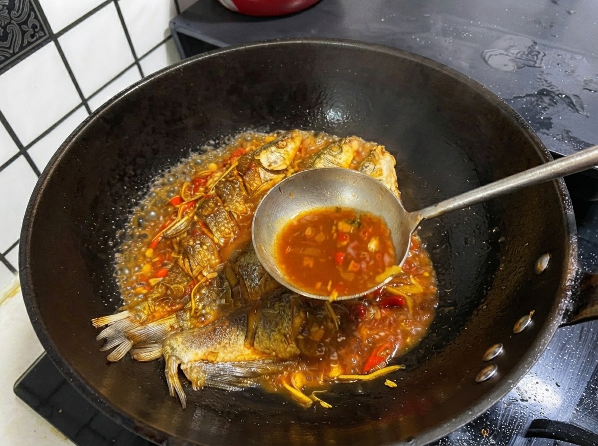 Un cucharón de metal sacando caldo oscuro y sabroso de una sartén que contiene pescado entero frito.