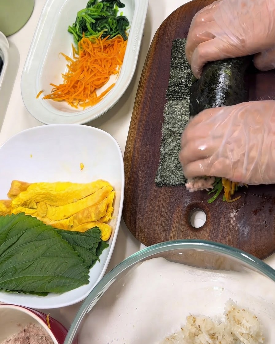Manos con guantes transparentes envolviendo ingredientes dentro de una hoja de nori en una tabla de cortar de madera.
