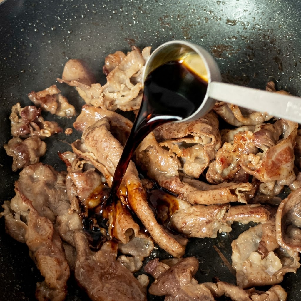 Una cuchara medidora vertiendo salsa de soja ligera oscura sobre tiras de carne cocida en una sartén.