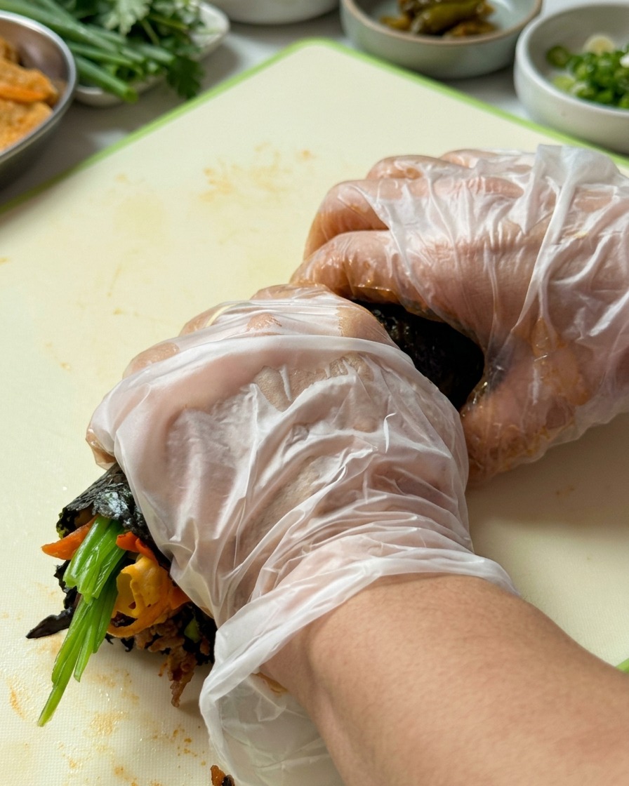 Manos con guantes enrollando firmemente el gimbap desde el borde inferior hacia arriba.