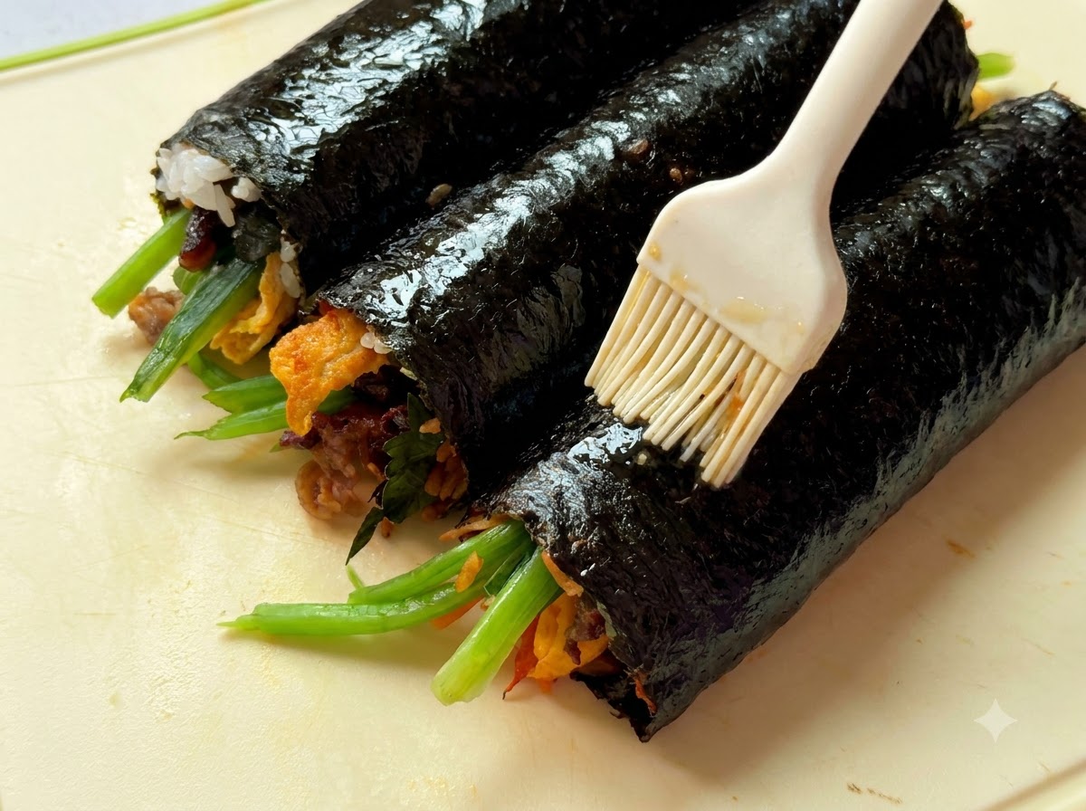 Vista cercana de aceite de sésamo siendo pincelado sobre tres rollos de gimbap colocados en una tabla de picar.