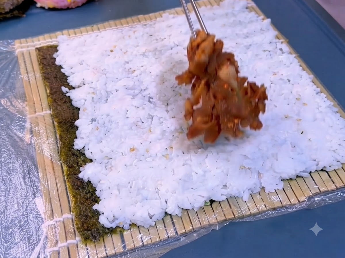 Una capa de arroz blanco cocido siendo extendida sobre una lámina de alga oscura en una esterilla de bambú.