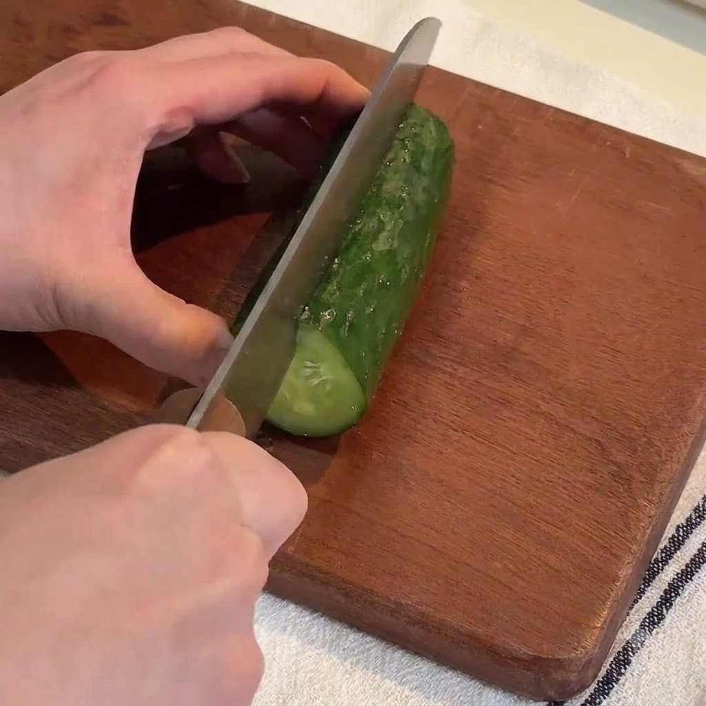 Manos usando un cuchillo grande para cortar un pepino verde fresco en tiras largas sobre una tabla de cortar de madera.