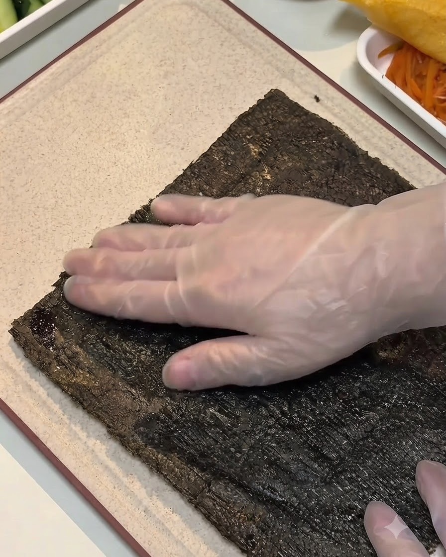 Una mano enguantada presionando hacia abajo el borde de una hoja de alga humedecida sobre una tabla de cortar.