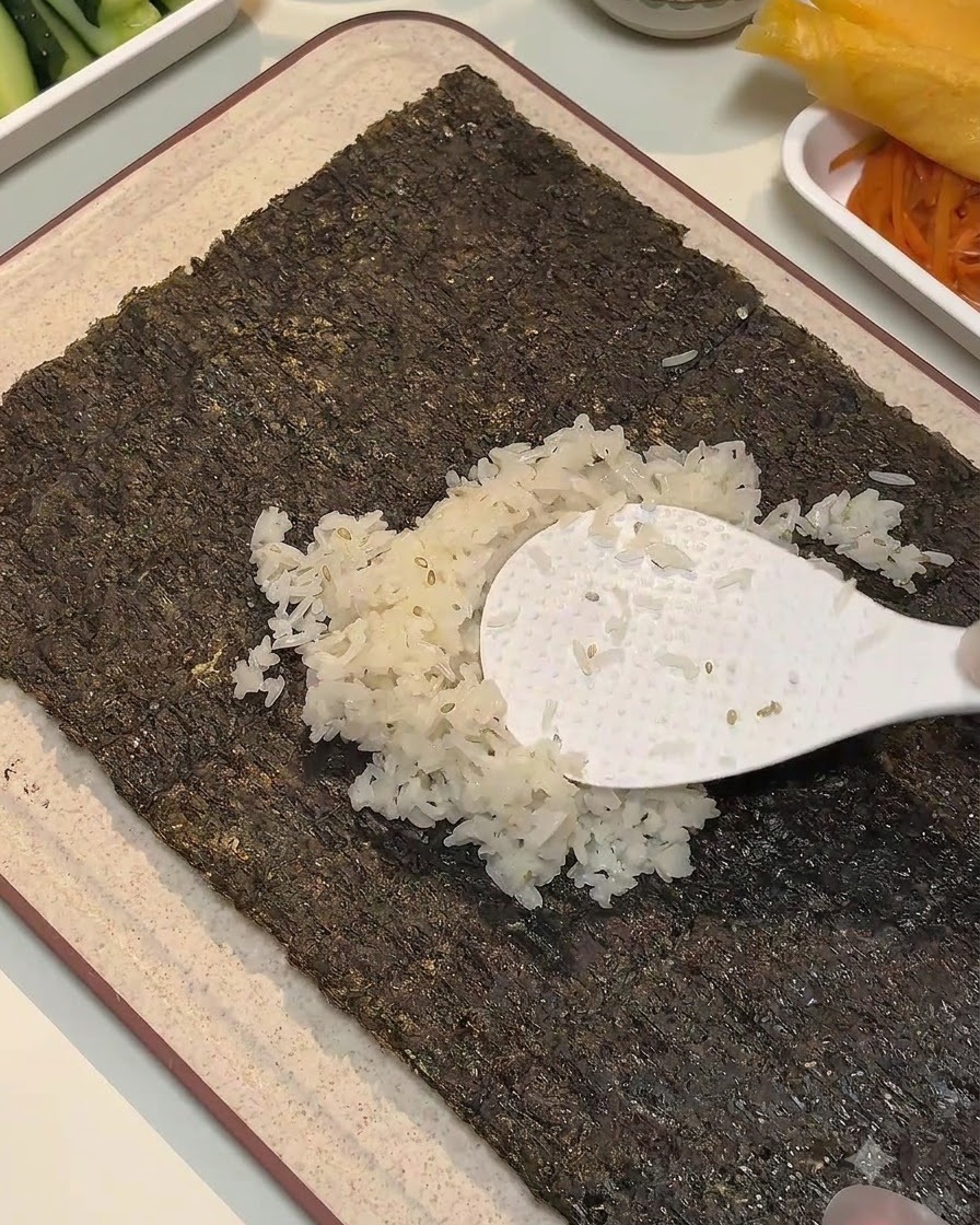 Una paleta de arroz blanca extendiendo arroz sazonado a lo largo del centro de una hoja de alga.