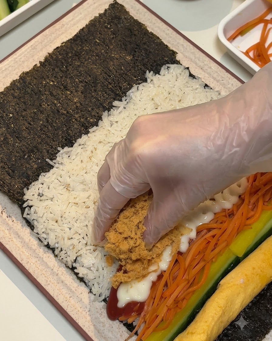 Una mano enguantada colocando carne desmenuzada marrón y esponjosa y tiras de jamón sobre los rellenos en la hoja de alga.