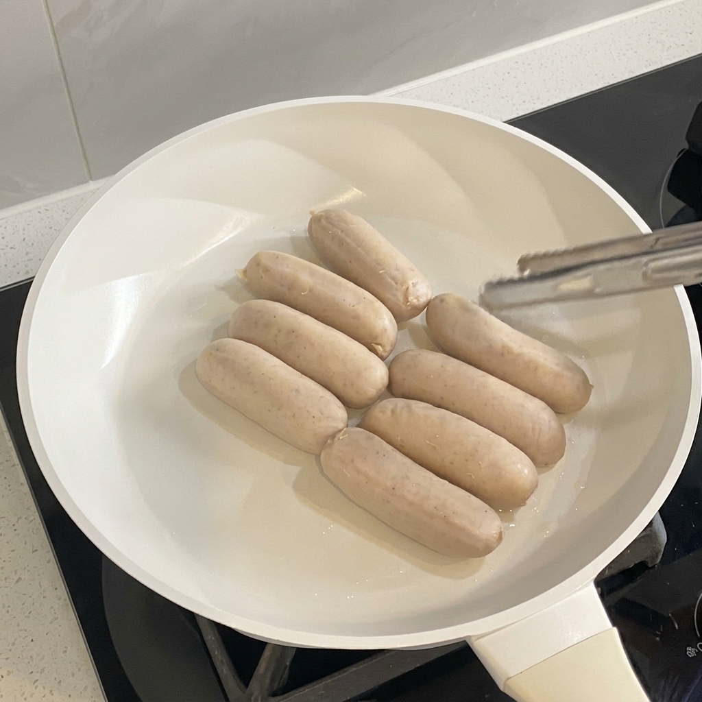 Salchichas cocinándose y dorándose de manera uniforme en una sartén blanca.