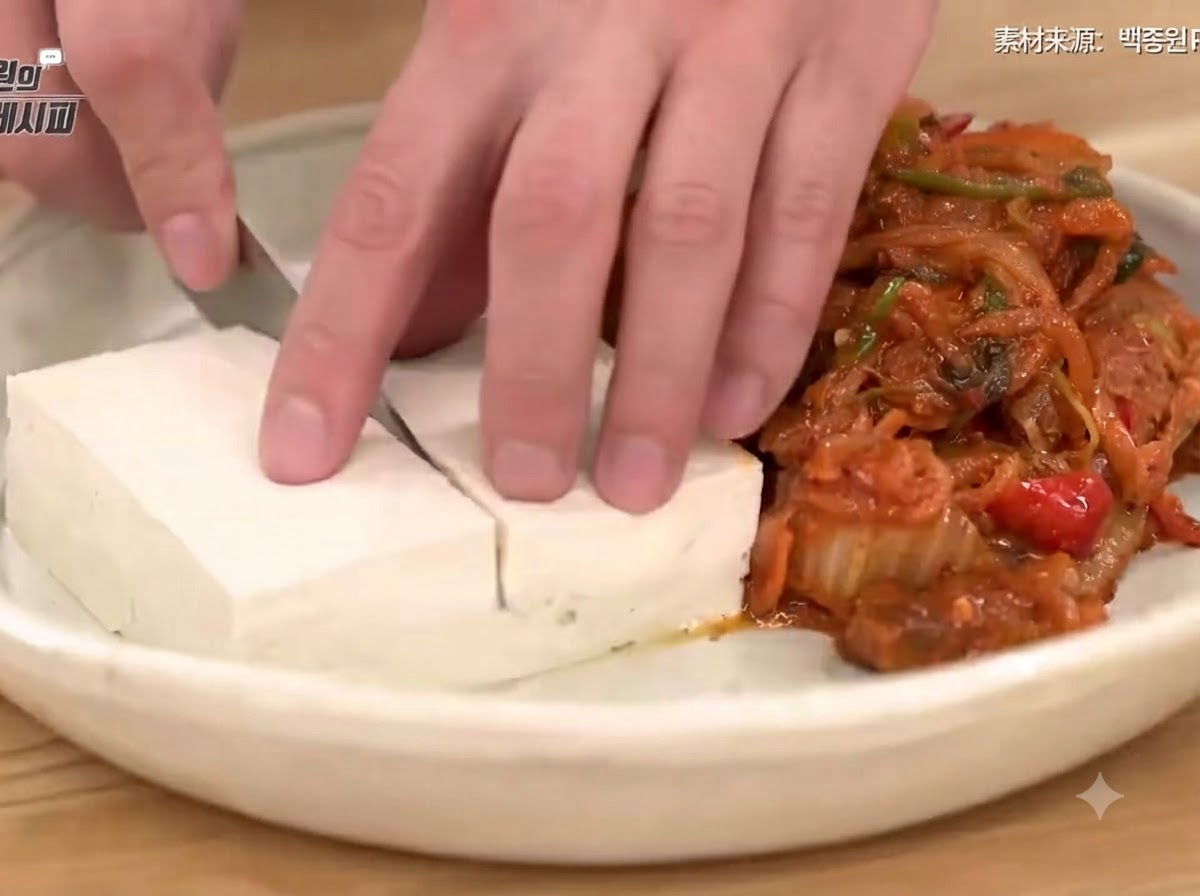 Una mano usando un cuchillo para cortar tofu escaldado ordenadamente en un plato junto a un montón de kimchi y cerdo salteados sabrosos.